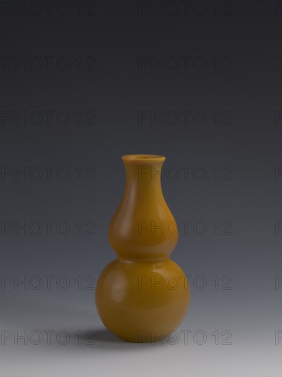 Vase, Chinese, Qing dynasty 1644–1911, 18th century. Double-gourd form vase made of yellow glass, cast and cut, opaque; stained glass; dimensions: height 16.5 cm, diameter 8 cm, weight 0.40 kg., A glossy mustard-yellow double-gourd shaped vase stands on a smooth grey surface against a dark grey gradient background, showing curved reflections and subtle highlights on its rounded body and narrow neck.