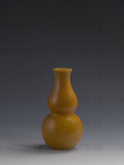 Vase, Chinese, Qing dynasty 1644–1911, 18th century. Double-gourd form vase of yellow glass, cast and cut, opaque. Dimensions: height 16.5 cm; diameter 8 cm; weight 0.40 kg. Subjects/attributes: gourds, stained glass., A smooth glossy double-gourd shaped vase in a warm yellow-orange color sits centered on a flat surface, showing subtle reflections and highlights on its curved surfaces; the background transitions from dark gray at the top to a lighter gray near the bottom, and a soft shadow in muted gray tones appears beneath and slightly to one side of the vase.