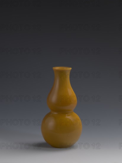 Vase, Chinese, Qing dynasty 1644–1911, 18th century. Double gourd-shaped vessel made of yellow glass, cast and cut, opaque; stained glass effect. Height 16.5 cm, diameter 8 cm, weight 0.40 kg., A glossy double-gourd shaped vase in a warm mustard yellow stands centered against a smooth gradient background that transitions from dark gray at the top to a lighter gray near the bottom, with a soft shadow on the surface beneath the vase.