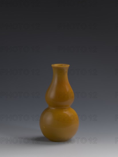 Vase, double gourds design, stained yellow glass, cast and cut, opaque; 18th century Qing dynasty, China; vessel; height 16.5 cm, diameter 8 cm, weight 0.40 kg., A smooth, glossy double-gourd shaped vase in a warm amber color sits centered on a gradient background that transitions from dark gray at the top to a lighter gray near the bottom, with a soft shadow beneath the vase and subtle highlights on its curved surfaces.