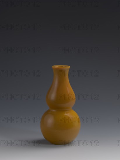 Vase, double gourd form; yellow glass, cast and cut, opaque; stained glass; gourds; Chinese; Qing dynasty 1644–1911, 18th century; vessel; height 16.5 cm; diameter 8 cm; weight 0.40 kg., A glossy gourd-shaped vase with a bulbous lower part and a narrower flared neck, rendered in a warm mustard yellow with highlights and subtle reflections, sits centered on a smooth surface that transitions from light gray near the vase to darker gray further away; the background above the vase is a deep charcoal gray that gradiently lightens toward the horizon, and the vase casts a soft shadow on the surface.