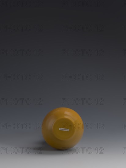 Vase, 18th century Qing dynasty China. Double gourd-shaped vessel of yellow glass, cast and cut, opaque; stained glass effect. Height 16.5 cm; diameter 8 cm; weight 0.40 kg., A single glossy round object in mustard yellow sits near the bottom center of the image against a smooth gradient background that transitions from dark gray at the top to lighter gray near the bottom, with a soft shadow beneath the object and faint reflections visible on its surface.