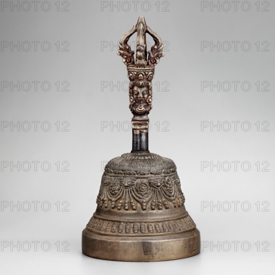Bell, 19th century, Tibet, ritual object vajra-related percussion idiophone, used in religious rites; material not specified; height 15 cm, diameter 7.3 cm., A single ornate metal bell sits upright against a neutral light gray background, showing a patina of brownish bronze and darker aged metal tones; the bell's handle rises into a decorative finial with sculpted faces and curved, openwork elements in a slightly lighter bronze color, while the shaft below the handle is darker and more blackened; the bell body displays a banded design with a textured top, a middle section of repeating draped and beaded motifs, and a lower band of small repeating oval decorations, all in varying shades of brown, bronze, and dark gray.