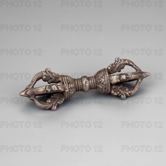 Vajra;, Himalayan, Tibetan Buddhist art; vajra ceremonial object; 18th century ; Tibet ; height 9.5 cm., The image shows a metallic object with an elongated symmetrical form resting on a flat surface; it has ornate sculpted sections with curved, leaflike and scrolllike decorative elements at both ends and a central banded, beaded area. The object appears to be made of a dark silvery metal with areas of darker patina and hints of brownish tones. The background and surface are a smooth, pale gray.