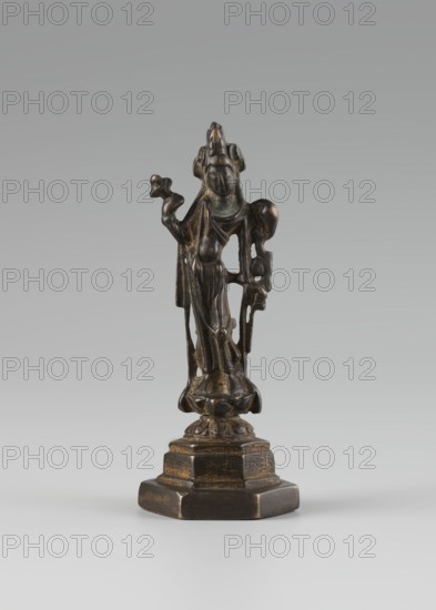 Bodhisattva;, Chinese culture; Chinese art; Chinese Buddhism; tutelary deities iconography; Asian culture; Buddhist art; Korean art; Goryeo 918–1392; 10th century ; Korea ; sculpture; figurine; height 11 cm, A small dark bronze figurine stands on a multi-tiered hexagonal base, showing a draped robed figure with a rounded headdress and stylized facial features; the figure’s garment falls in vertical folds and there are openwork areas along the sides. The figure’s arms are raised, one holding a small modeled object and the other bending to touch a curved element, with additional small ornamental details at the shoulders and hips. The surface shows areas of lighter brown and greenish patina against a predominantly dark bronze color, and the background is a smooth gradient of light gray.