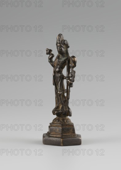 Bodhisattva, Chinese culture, Chinese Buddhism, tutelary deities iconography, Asian culture, Buddhist art, Korean art, Goryeo 918–1392, 10th century, Korea place of origin ?, sculpture, figurine, height 11 cm., A small dark bronze-colored standing figurine on a stepped hexagonal base, with a weathered surface showing areas of brownish-gold highlights and darker greenish-brown patina; the figure is elongated and robed, with a raised arm holding a small rounded object and the other arm bent at the side, a rounded head with a stylized headdress or hair detail, draped clothing with visible folds, and an overall aged metallic texture against a neutral gray background.