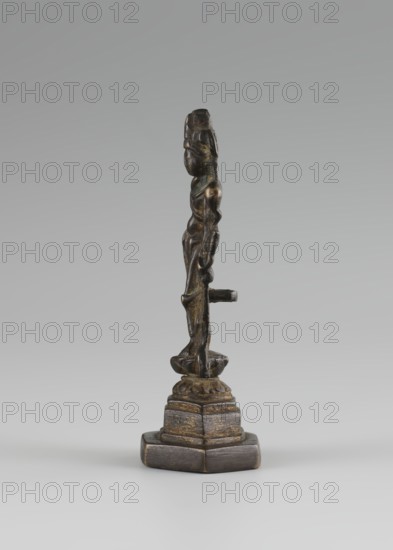 Bodhisattva, Chinese culture, Chinese art, Chinese Buddhism, tutelary deities iconography, Asian culture, Buddhist art, Korean art, Goryeo 918–1392, 10th century, Korea place of origin ?, sculpture, figurine, height 11 cm, A slender vertical sculpture of a standing figure on a multi-tiered base, rendered in dark bronze tones with areas of brown and muted gold highlights, set against a pale gray background; the figure shows a simplified face and elongated limbs with a small horizontal protrusion at waist level, the base appears polygonal and textured with darker brown and golden wear, and the overall palette includes dark bronze, brown, muted gold, and gray.