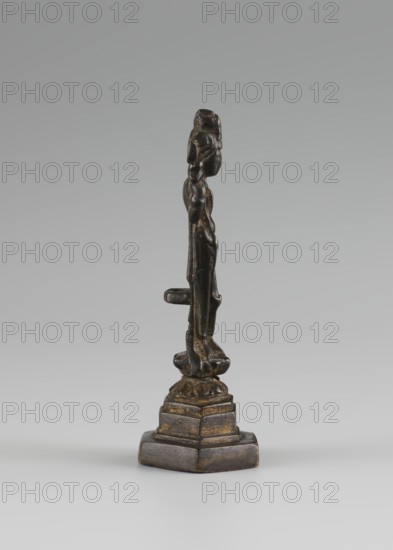 Bodhisattva, Chinese; Chinese art; Chinese Buddhism; tutelary deities iconography; Asian; Buddhist art; Korean art; Goryeo 918–1392; 10th century : Korea place of origin?; sculpture, figurine; height 11 cm, A narrow dark metallic figurine stands on a stepped base against a pale gray background; the figure has a vertical elongated form with a rounded head, a curved torso, and a few protruding elements along its side, displaying worn areas where a golden-brown metal appears beneath the darker surface, and the base shows layers of dark gray and bronze tones.