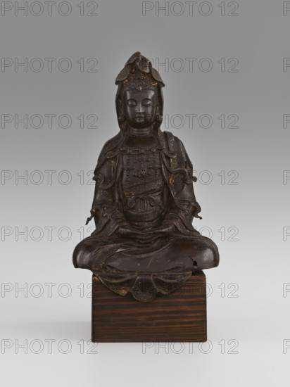 Guanyin, Avalokiteshvara iconography, Chinese Buddhism, Chinese art, Chinese culture, goddesses, bodhisattva iconography, mercy, 16th/17th century, China, sculpture, figurine, height 28.0 cm, A dark brown sculpted figure seated cross-legged on a rectangular wooden block, with detailed draped robes and layered adornments, a rounded headdress with petal-like elements, a calm closed-eyed facial expression, subtle worn lighter brown and golden highlights on raised surfaces, and the wooden base showing striped brown grain against a neutral gray background.
