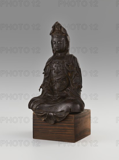 Guanyin, Avalokiteshvara iconography, Chinese Buddhism, Chinese art, Chinese culture, goddesses, bodhisattva iconography, mercy, 16th/17th century, China, sculpture, figurine, height 28.0 cm, A dark brown statue of a seated figure with a serene facial expression rests atop a wooden block base; the figure wears a detailed headdress and layered robes with carved decorative patterns and flowing folds, showing some worn areas and small patches of lighter brown or golden discoloration; the wooden base displays a natural wood grain in shades of brown; the background is a smooth gradient of gray transitioning from darker at the top to lighter toward the bottom.