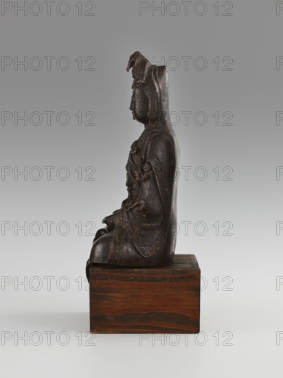 Guanyin, Avalokiteshvara iconography, Chinese Buddhism, Chinese art, Chinese culture, goddesses, bodhisattva iconography, mercy; 16th/17th century, China; sculpture, figurine; height 28.0 cm; creator, provenance: purchase., A dark brown sculpted figure seated sideways on a rectangular wooden base, with a smooth rounded back and detailed carved drapery and ornamental patterns along the garment, showing traces of lighter brown or gold highlights in the carved areas; the figure wears a hood or headdress with layered elements and a calm profile with closed eyes, all set against a neutral gray gradient background with a pale gray surface beneath the base.