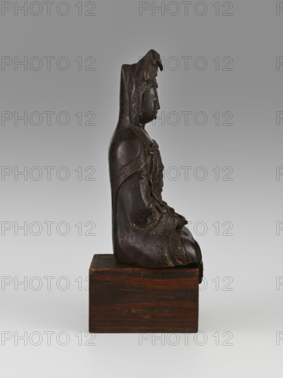 Guanyin, Avalokiteshvara iconography, Chinese Buddhism, Chinese art, Chinese culture, goddesses, bodhisattva iconography, mercy, 16th–17th century, China, sculpture, figurine, height 28.0 cm, A dark brown sculpted figure seated on a wooden block, shown in profile facing right, with a hooded or draped garment and detailed carved patterns on the clothing, a rounded face with closed or relaxed eyes, hands resting in the lap, and areas of subtle lighter brown and goldish highlights on the carved details; the wooden block is a warm medium brown with visible grain and darker streaks, and the background is a smooth gradient of light gray to a slightly darker gray.