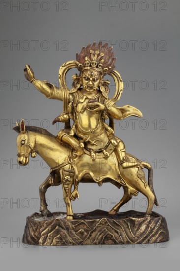 Palden Lhamo; unknown creator; sculptor; purchased provenance; Tibetan Buddhist art; Himalayan art; Himalayan culture; dharmapala iconography; mules; human skins; tiaras; skulls; 18th century, Tibet; sculpture figurine; height 16.7 cm; weight 1.11 kg, A gilded figurine of a fierce deity riding a horse stands on a textured base; the deity has an ornate headdress with flame-like elements that are reddish-brown and gold, a stern facial expression, and elaborate jewelry and drapery in gold tones, with arms posed in dynamic gestures and a looping decorative sash behind the shoulders. The horse is gold with darker accents on its mane, tail, and hooves and carries the rider with a decorative saddle and harness. The base is dark brown with gold highlights and a carved, wave-like pattern. The background is a neutral gray.