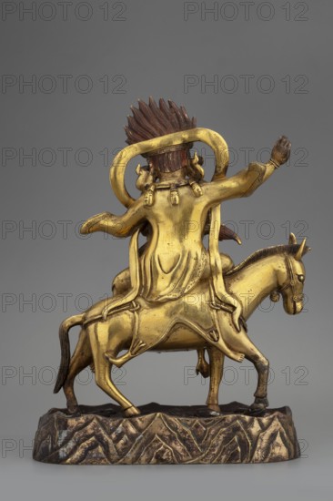 Palden Lhamo, unknown sculptor, Tibetan Buddhist art, Himalayan art, Himalayan culture, dharmapala iconography, mules, skins human, tiaras, skulls, 18th century, Tibet, sculpture, figurine, height 16.7 cm, weight 1.11 kg, A gilded metal figure of a person seated on a horse, shown from the back, with a flowing robe and an ornate sash draped over shoulders and down the rider’s back; the rider has a tall, spiked hairpiece in a darker brown tone and a rounded halo-like element behind the head. The horse and rider are both gold in color with darker brown accents on the hairpiece, tail, hooves, and base. The horse stands on an irregular textured base with a mottled gold and dark brown surface that suggests a rocky ground. The rider’s raised arm and the sculpted folds of the robe are clearly visible, as are small decorative details on the rider’s sleeves and the horse’s bridle. The background is a smooth, neutral gray.
