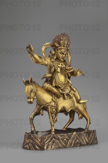 Palden Lhamo, unknown sculptor, Tibetan Buddhist art, Himalayan art, Himalayan culture, dharmapala iconography, mules, human skins, tiaras, skulls, 18th century, Tibet, sculpture, figurine, height 16.7 cm, weight 1.11 kg, A gilded figure with a golden-yellow finish sits astride a horse on a low textured base; the figure wears an ornate headdress with dark reddish-brown accents and multiple small decorative elements, a fierce facial expression, and layered necklaces and drapery in matching gold tones, with one arm raised and the other bent toward the chest holding a small rounded object; the horse is also gold with darker shading on its ears and mane, decorative harnessing and a draped saddle, and both figure and horse rest on a mottled brown and gold base against a neutral gray background.