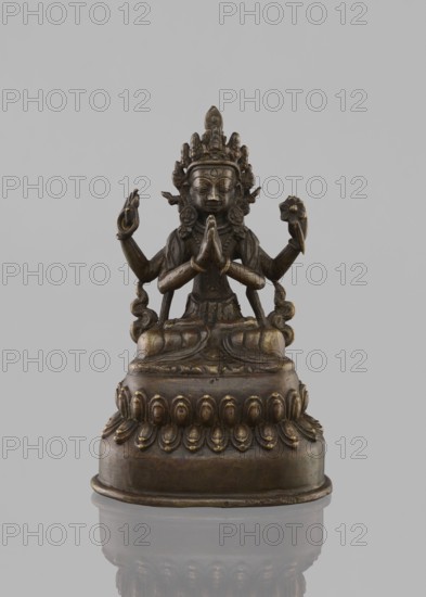 Bodhisattva Avalokiteshvara Sadaskari, 18th–19th century, Nepal. Sculpture/figurine/cult object, height 17.5 cm. Bodhisattva Sadaksari sPyan-sras-gzigs in dhanasana on a double lotus throne; one pair of hands folded in namaskara mudra, third hand holding a mala and padma. Wearing dhoti, billowing scarf and jewelry; includes mala prayer beads, lotus throne, statuette form, chingon, amitabha? drop-shaped urn. Buddhist/Nepalese art, bodhisattva iconography., A small bronze-colored statue of a seated figure on a rounded ornate base, shown against a plain light gray background. The figure has a detailed headdress and an intricate necklace, with a calm facial expression and closed eyes. The figure sits cross-legged with hands held together at chest level and additional arms extended to the sides holding small objects, and there are decorative elements and flowing drapery around the torso and limbs. The base features a row of petal-like motifs and a reflective surface beneath the statue shows...