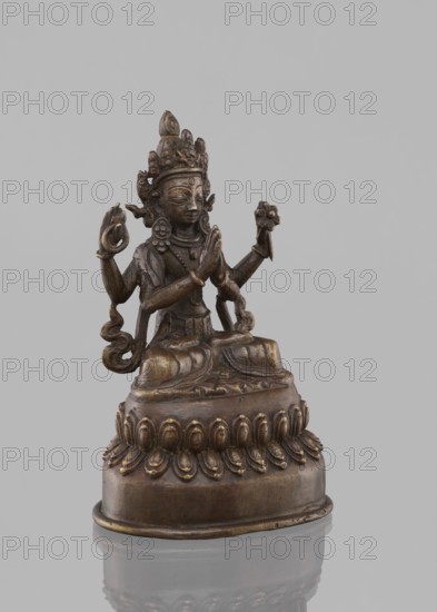 Bodhisattva Avalokiteshvara Sadaskari, 18th–19th century, Nepal. Sculpture/figurine cult object, height 17.5 cm. Bodhisattva Sadaksari sPyan-sras-gzigs — in dhanasana on a double lotus throne; one pair of hands folded in namaskara mudra, a third hand holding a mala and padma; wearing a dhoti, billowing scarf, jewelry; includes mala prayer beads; possibly associated with Amitabha; drop-shaped urn; statuette, Nepalese Buddhist art., A small sculpted figure sits on a rounded base with a decorative lotus-like rim, rendered in brownish bronze tones with areas of darker and lighter patina; the figure has multiple arms, hands positioned in various gestures, and holds a small floral form in one hand, with ornate jewelry and a tall, intricate crown atop the head; flowing drapery and curled ornamental elements extend from the shoulders and arms, and the figure gazes forward with a calm facial expression; the background is a smooth, light gray.