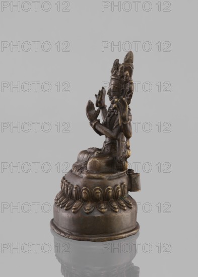 Bodhisattva Avalokiteshvara Sadaskari, 18th–19th century, Nepal. Sculpture/figurine cult object, height 17.5 cm. Bodhisattva Sadaksari sPyan-sras-gzigs in dhanasana on a double lotus throne; one pair of hands folded in namaskara mudra, a third hand holding a mala and padma; wearing a dhoti, billowing scarf, jewelry; includes mala prayer beads, lotus throne, possible amitabha or drop-shaped urn. Style: Nepalese Buddhist art, statuette., A small bronze statue of a seated figure on a decorative round base is pictured against a light gray background, showing a side view. The figure has a detailed headdress and multiple layered adornments, with hands raised in a gesture and draped garments visible. The base features a lotus-like petal motif around its upper edge and a rounded lower platform. Visible colors include bronze tones, dark brown, muted gold highlights, and the light gray of the background.