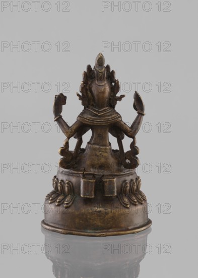 Bodhisattva Avalokiteshvara Sadaskari, Bodhisattva Sadaksari sPyan-sras-gzigs in dhanasana on a double lotus supported by a base; one pair of hands folded in namaskara mudra, third hands holding a mala and padma; wearing dhoti, billowing scarf and jewelry; chignon, possibly Amitabha or drop-shaped urna; mala prayer beads; lotus throne; statuette/figurine cult object; Nepalese Buddhist art; 18th–19th century; sculpture; height 17.5 cm, A small metal statue viewed from the back, with a dark bronze surface showing areas of lighter brown and subtle greenish patina, seated on a rounded pedestal decorated with petal-like reliefs; the figure has a multi-tiered headdress and a neck collar, with a group of flowing ornamental elements extending from the shoulders and a few raised arms bent at the elbows, the overall silhouette showing worn edges and polished highlights against a neutral gray background.