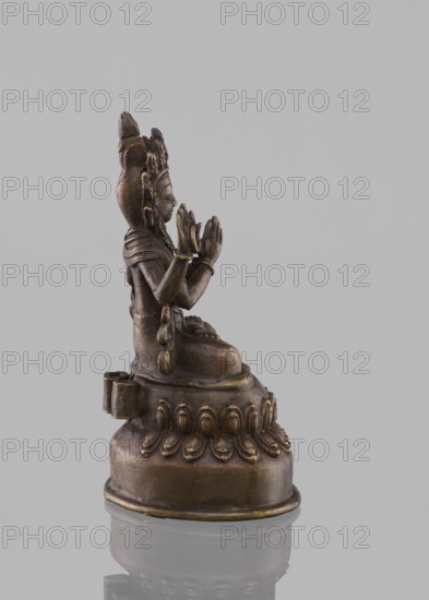 Bodhisattva Avalokiteshvara Sadaskari, Nepalese Buddhist sculpture, 18th–19th century. Statuette figurine, cult object of Bodhisattva Sadaksari sPyan-sras-gzigs seated in dhanasana on a double lotus lotus throne. One pair of hands folded in namaskara mudra; a third hand holds a mala and padma; includes mala prayer beads, dhoti, billowing scarf, jewelry, chignon, possibly Amitabha or a drop-shaped urn. Height 17.5 cm., A small bronze-colored statue of a seated figure on an ornate rounded base is shown in profile against a pale gray background; the figure has a tall headdress, detailed facial features, layered garments, and hands held together in front of the chest, with decorative elements and dangling ornaments along the side of the torso, and the base displays a row of petal-shaped motifs and a darker bronze patina.