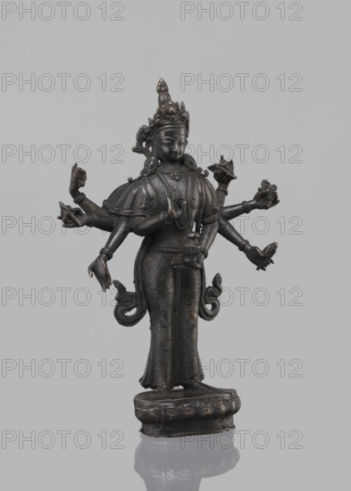 Avalokiteshvara Bodhisattva, Tibetan Buddhist art, Buddhist art, Himalayan art, bodhisattva iconographic, lotus throne iconographic, 17th century, Nepal, sculpture, figurine, cult object, height 24.5 cm, A dark metal statue of a multi-armed figure standing on an ornate round base against a plain light gray background; the figure wears an elaborate headdress and jewelry, has a serene facial expression, and is dressed in a long patterned garment with flowing decorative elements at the hips; several arms extend from the torso in various gestures, some holding small sculpted objects, and the metal shows subtle highlights of worn lighter tones on raised areas.