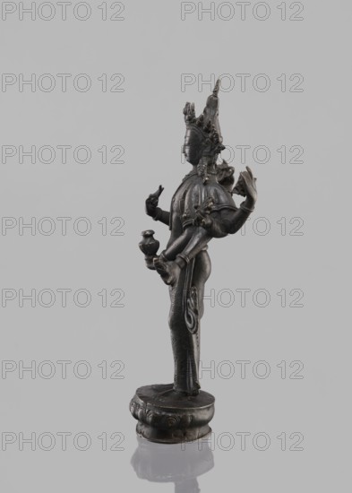 Avalokiteshvara Bodhisattva, Tibetan Buddhist art, Buddhist art, Himalayan art, bodhisattva iconographic, lotus throne iconographic, 17th century, Nepal, sculpture, figurine, cult object, height 24.5 cm, A dark metallic statue stands on a rounded base with decorative edges, shown in profile against a light gray background. The figure has a tall ornate headdress and detailed facial features, with visible earrings and an intricate necklace. Several arms extend from the torso, some holding small objects shaped like vessels or symbolic items, and the hands display varied gestures. The clothing and body are decorated with fine engraved patterns and flowing drapery elements, and the surface shows subtle highlights of lighter metallic tones against the predominant dark metal. The base and lower part of the statue reflect faintly on the smooth surface below, and the overall colors visible are dark gray, light gray, and small areas of slightly warmer metallic sheen.