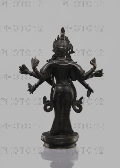 Avalokiteshvara Bodhisattva, Tibetan Buddhist art, Buddhist art, Himalayan art, bodhisattva iconographic, lotus throne iconographic, 17th century, Nepal, sculpture, figurine, cult object, height 24.5 cm, A dark bronze-colored statue viewed from the back, standing on a rounded base with a reflective surface beneath. The figure has a smooth, elongated silhouette with a flared lower garment and a narrow waist, and wears an ornate headdress with pointed and layered elements. Several arms extend outward from the torso in different directions, some holding indistinct small objects, and the hands show varied gestures. There are decorative looping extensions at the hips and subtle raised details on the shoulders and back. The background is a plain light gray, and the visible colors are dark bronze, black, and shades of gray.