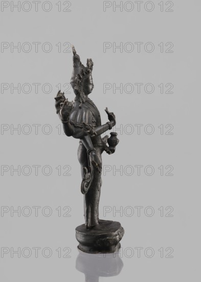 Avalokiteshvara Bodhisattva, Tibetan Buddhist art, Buddhist art, Himalayan art, bodhisattva iconographic, lotus throne iconographic, 17th century, Nepal, sculpture, figurine, cult object, height 24.5 cm, A dark metallic statue of a standing figure on a rounded base against a plain light gray background, showing a side view of the figure with a tall ornate headdress and detailed facial features, multiple arms extending in different directions holding small objects, intricately carved garments and jewelry with visible decorative patterns, the metal surface appearing dark gray with subtle highlights of lighter gray and small areas of worn bronze or golden tones, a reflective surface beneath the base showing a muted mirror of the statue.