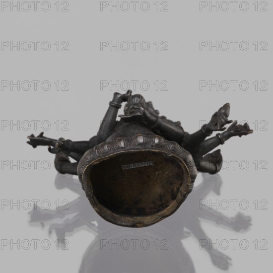 Avalokiteshvara Bodhisattva, Tibetan Buddhist art, Buddhist art, Himalayan art, bodhisattva iconographic, lotus throne iconographic, 17th century, Nepal, sculpture, figurine, cult object, height 24.5 cm, A small dark metal sculpture is shown lying on its back on a reflective light gray surface, revealing a hollow base with a rim that has a scalloped edge; the sculpture has multiple outstretched arms and hands with detailed fingers, a central torso with ornamental drapery and raised decorative elements, and a head with carved facial features and hair elements. The visible colors are dark bronze, muted brown inside the hollow base, and light gray from the background and reflection.