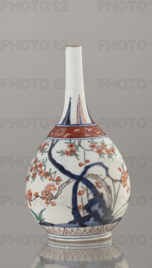 Bottle, Japanese, Edo 1603–1868, early 18th century. Porcelain vessel sake bottle, height 22.1 cm, diameter 12 cm. Decorated in the kakiemon style with a motif of the three friends of winter bamboo, plum, and pine; rock. Also features two borders with plant motifs and floral patterns on the neck., A tall, narrow-necked ceramic vase with a bulbous body sits against a neutral gray background; the vase is primarily white with painted decoration in blue, red, green, and orange. A band of red with floral motifs encircles the upper shoulder, and below it a dark blue, twisting tree-like form extends across the body, surrounded by clusters of orange and red blossoms and green leaves. Additional blue and red linear patterns decorate the base and neck, and areas of the glaze show fine crackling. The vase rests on a reflective surface that mirrors its base and some of the painted patterns.