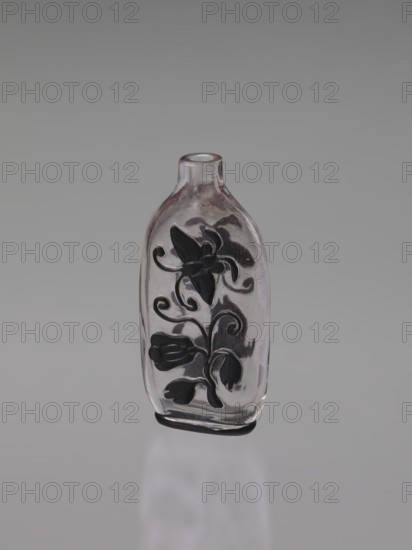 Flask - snuffbox; cylindrical neck; layered transparent glass with a black top layer; decoration cut in the top layer depicting flowers, lion, bottles, and butterflies; without stopper; 19th century, China; Qing 1644–1911; height 7.6 cm, width 3.8 cm., A small clear glass bottle stands upright against a neutral gray background; the bottle has a short neck and an oval body with a decorative raised floral design in dark color on its front, showing stylized petals, buds, and curling stems; the glass shows subtle reflections and a slight pinkish tint in places, and the dark decorative elements appear black or very dark gray; the surface beneath the bottle creates a soft pale reflection.