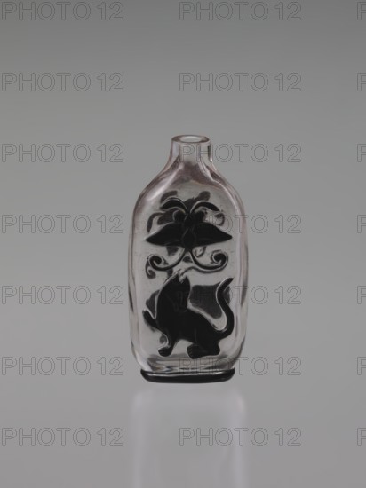 Flask snuffbox, 19th century, China, Qing 1644–1911. Layered transparent glass with top black layer; decoration cut in the top layer depicting flowers, lion, bottles, and butterflies. Cylindrical neck; without stopper. Vessel, height 7.6 cm, width 3.8 cm., A small clear glass bottle with a rounded rectangular shape and a short neck stands upright against a smooth gray background; the bottle contains black decorative silhouettes on its front, including a stylized floral or leafy motif near the upper portion and a playful animal figure resembling a cat with a raised paw near the lower portion, and the glass shows subtle reflections and slight pinkish tones around the neck while the base rests on a reflective surface that creates a soft mirrored shadow; visible colors include clear/transparent glass, black decoration, gray background, and a hint of pink.
