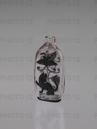 Flask-snuffbox, ; motif: flowers, lion, bottles, butterflies; Chinese art, Chinese culture; Qing dynasty 1644–1911; cylindrical neck; layered transparent glass with black top layer; decoration cut in the top layer; without stopper; 19th century; China; vessel; height 7.6 cm, width 3.8 cm, A small transparent glass bottle with a narrow neck and rounded shoulders stands against a neutral gray background. The bottle has black raised decorations on its front, including a stylized flower near the top with curling stems and a silhouette of a cat with a curved tail toward the lower part. The glass shows subtle reflections and a few pale pinkish tints along its edges, and there is a thin dark band at the base. The overall visible colors are clear/transparent glass, black decoration, pale pink highlights, and gray from the background.