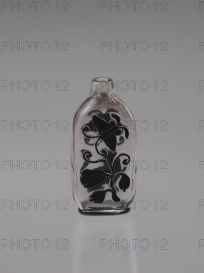 Flask-snuffbox, cylindrical-necked layered glass with transparent body and black top layer; decoration cut in the top layer depicting flowers, lion, bottles, and butterflies. Without stopper. 19th century, Qing dynasty 1644–1911, China. Height 7.6 cm; width 3.8 cm., ., A small clear glass bottle stands upright against a neutral gray background, with a smooth rounded shoulder and a short narrow neck; a decorative dark floral motif is visible on its front, showing stylized petals, leaves, and curling stems in black or very dark gray; the glass has subtle reflections and a few faint surface imperfections, and the base shows a thin dark ring; overall the visible colors are clear/transparent, gray, and black.