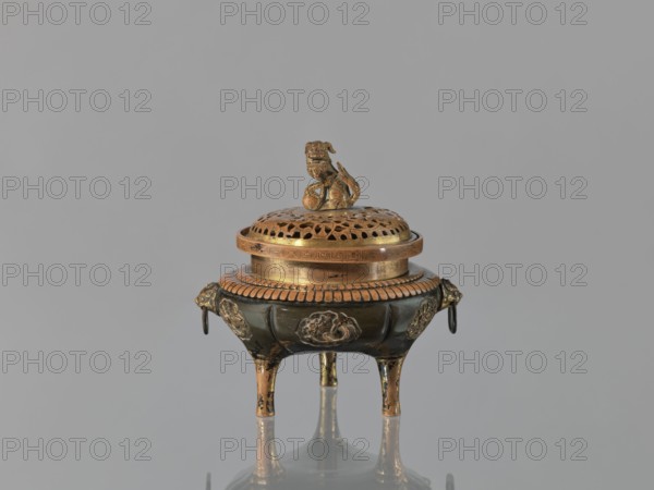 Incense burner koro of the ding type with a cover. Incense burner with two handles knobs, on three legs. Openwork cover decorated with a floral tendril and topped with a komainu figure mythical beast. Meiji period 2nd half of the 19th century, 1868–1912. Japan. Height 13.8 cm; diameter 12.9 cm., An ornate metal censer sits centered against a gray background, featuring a domed perforated lid topped by a small sculpted figurine; the body rests on three slender legs and has decorative appliqués and ring handles on its sides. The visible colors include gold, dark bronze, black, and hints of brown, with the gold appearing on the lid, figurine, rim, decorative details, and leg accents while darker bronze and black color the main body and recessed areas. The surface displays polished highlights and areas of darker patina, and a soft reflection of the object appears on the surface beneath it.