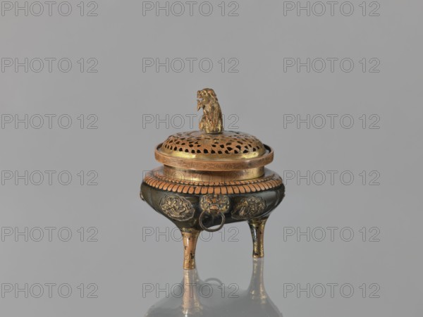 Incense burner kôro of the ding type with a cover. Incense burner with two handles knobs, on three legs. Openwork cover decorated with a floral tendril and topped with a komainu figure mythical beast. Meiji period, 2nd half of the 19th century 1868–1912. Japan. Bronze, height 13.8 cm, diameter 12.9 cm., The image shows a small ornate tripod vessel with a perforated lid topped by a sculpted animal figure; the vessel sits on a reflective surface against a plain background. The object displays metallic colors including gold, bronze, and dark brown, with a few areas of lighter worn metal; the lid has a patterned perforation and a textured rim, the body features decorative raised medallions and a ring hanging from one side, and the three tapered legs show wear and metallic sheen.