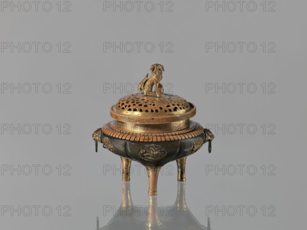 Incense burner koro of the ding type with a cover. Incense burner with two handles knobs, on three legs. Openwork cover decorated with a floral tendril and topped with a komainu figure mythical beast. Meiji period, 2nd half of the 19th century 1868–1912. Japan. Height 13.8 cm; diameter 12.9 cm., floral pattern; ding vessel form; incense burner function., An ornate metal incense burner sits on three short legs on a reflective surface, with a perforated domed lid topped by a small animal-shaped finial; the burner shows a mix of metallic gold and dark brown tones, with decorative raised motifs and a ridged band around the lid, and a few dangling ring ornaments at its sides, all set against a plain light gray background.
