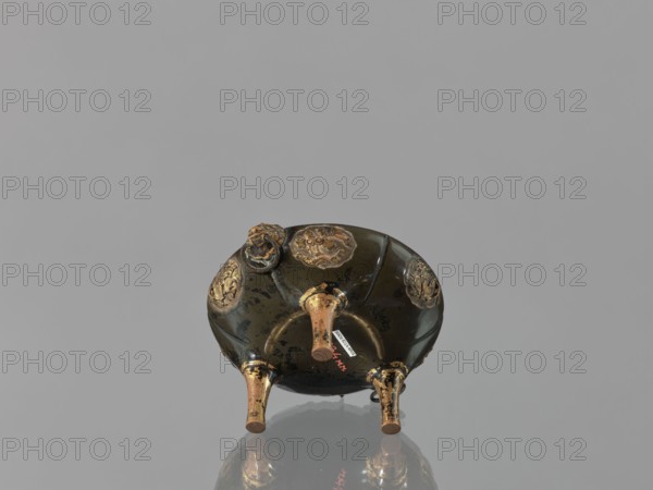 Incense burner koro of the ding type with a cover. Incense burner with two handles knobs, on three legs. Openwork cover decorated with a floral tendril and topped with a komainu figure mythical beast. Meiji period 2nd half of the 19th century, Japan. Bronze? material unspecified. Height 13.8 cm, diameter 12.9 cm., A small rounded object with a dark greenish-black surface and areas of worn gold detailing sits on a reflective gray surface; it rests on three short conical legs that are gold and show signs of wear, and the underside displays several raised decorative medallions in gold with textured patterns, along with a small protruding knob near the edge.