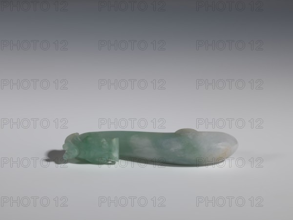 Belt clasp, purchase provenance. Chinese, Qing 1644–1911. Green-gray jadeite, semi-transparent with slight colloid clusters. Full-plastic carved head with dragons iconography. Smooth handle. Function: belt buckle for men's clothing, ornament. 17th–18th century. Dimensions: height 2.0 cm, width 9.1 cm, depth 1.7 cm., A smooth, elongated carved object lies horizontally on a light surface against a gradient gray background; the object is primarily green with areas of pale white and subtle brownish discoloration, and one end features a carved decorative form with rounded protrusions while the other end is rounded and smooth, casting a soft shadow beneath.