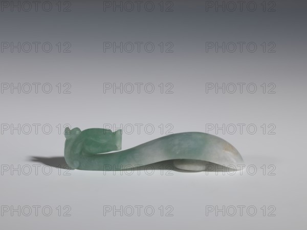 Belt clasp, Chinese, dragons iconography, Qing 1644–1911, men's clothing/belts/ornaments, smooth handle. Green-gray jadeite, semi-transparent with slight colloid clusters. Fully carved in-the-round head. 17th–18th century, China. Belt buckle; H 2.0 cm, W 9.1 cm, D 1.7 cm., A smooth, curved object made of translucent green and pale blue stone rests on a neutral surface, forming an elongated S-shape with one end carved into a rounded, stylized animal head and the other tapering to a gently rounded point; the stone shows subtle variations in color from deeper green to lighter blue-gray with faint cloudy areas and a small patch of warmer discoloration, and a small circular stone support is visible beneath the midsection, casting a soft shadow on the surface.