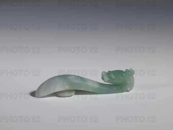 Belt clasp, Chinese, dragons iconography, Qing 1644–1911, men's clothing ornament; smooth handle. Green-gray jadeite, semi-transparent slight colloid clusters. Full-plastic carved head. 17th–18th century, China. Belt buckle; H 2.0 cm, W 9.1 cm, D 1.7 cm., A smooth carved object made of translucent green and pale grey stone rests on a neutral surface, the stone showing gradients from light green to milky grey; the form is elongated and curved with a rounded broad end tapering to a sculpted head-like end featuring rounded protrusions and subtle carved details, and a small circular support under the curved middle; the background is a gradient from light grey at the bottom to darker grey at the top.