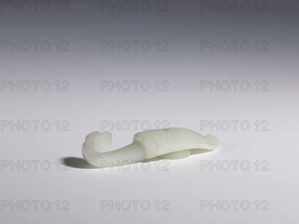 Belt clasp, Chinese, Chinese art, cicadas, lizards zool., Qing 1644–1911. Stem: stylized cicada with a geometric meander motif. Milky-white jadeite, slightly translucent visible clusters of colloids. Head carved in full relief, flat relief. 17th–18th century, China. Belt buckle. Height 1.8 cm, width 8.0 cm, depth 2.1 cm., A smooth pale white carved object resembling an elongated animal form rests on a light surface, with a curved tail or neck ending in a rounded head at one end and a domed, patterned shell or back at the other; the carving shows shallow raised geometric and circular motifs, smooth rounded contours, and casts a soft gray shadow on the surface beneath. The visible colors are white, pale off-white, light gray, and darker gray in the background gradient.