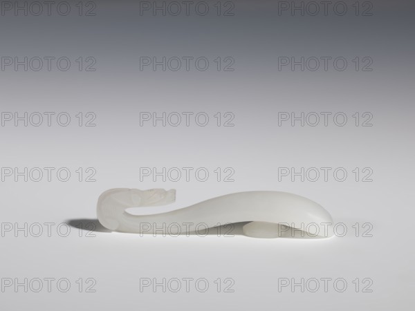 Belt clasp, Chinese, Chinese art, dragons iconography, Qing 1644–1911, handle devoid of engraved decoration, polished smoothly. Milky-white jadeite, slightly translucent. Full-plastic carved head, engraved, 18th–19th century, China, belt buckle; height 1.5 cm, width 8.8 cm, depth 1.7 cm., A smooth, curved white object resembling a stylized animal form rests on a pale surface, with a carved, detailed head at one end and a gently arching body that tapers toward the other end; the object casts a soft shadow and sits on a small round white support, and the background transitions from light gray near the surface to a darker gray toward the top.
