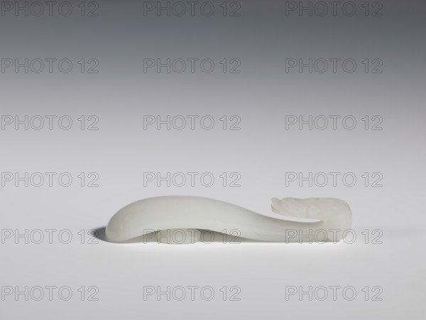 Belt clasp, Chinese, dragons iconography, Qing 1644–1911, handle devoid of engraved decoration, polished smoothly. Milky-white jadeite, slightly translucent. Full-plastic carved head, engraved, 18th–19th century, China, belt buckle, height 1.5 cm, width 8.8 cm, depth 1.7 cm, A smooth, curved pale object with a translucent white appearance rests on a flat surface, showing a rounded, thicker midsection and a tapered end that curls upward into a more detailed carved shape; the background is a gradient from dark gray at the top to light gray near the surface, and the object casts a soft shadow on the surface.