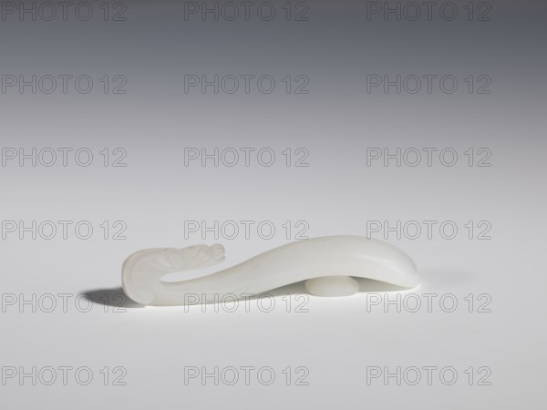 Belt clasp, Chinese, dragons iconography, Qing 1644–1911, handle devoid of engraved decoration, polished smoothly. Milky-white jadeite, slightly translucent. Full-plastic carved head, engraved, 18th–19th century, China, belt buckle, height 1.5 cm, width 8.8 cm, depth 1.7 cm, A smooth, curved object with an elongated, undulating shape rests on a flat surface, appearing to be made of a translucent white material; one end tapers into a carved, slightly ornate form while the other end rises into a rounded, dome-like curve that is supported by a small circular base, casting soft shadows on the surface beneath; the background transitions from light gray near the surface to darker gray toward the top, and visible colors are shades of white and gray.
