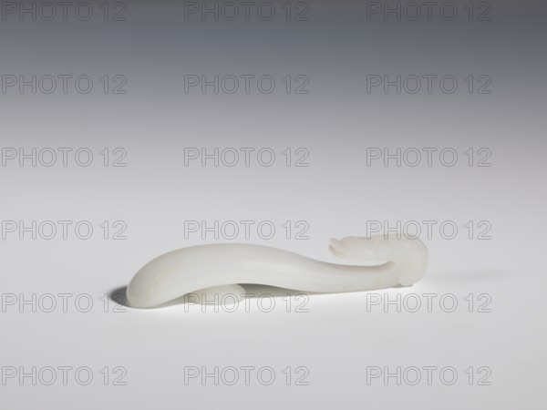 Belt clasp, Chinese, dragons iconography, Qing 1644–1911, handle devoid of engraved decoration, polished smoothly. Milky-white jadeite, slightly translucent. Full-plastic carved head, engraved, 18th–19th century, China, belt buckle; height 1.5 cm, width 8.8 cm, depth 1.7 cm., A smooth, curved object made of a translucent white material rests horizontally on a flat surface, casting a soft shadow beneath it; the object tapers at one end and has a slightly bulbous, rounded opposite end with subtle carved ridges and contours; the background transitions from medium gray at the top to pale gray near the surface, and the visible colors are white, gray, and faint cream tones on the object.
