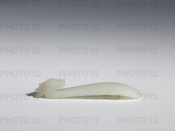 Belt clasp, purchase provenance; Chinese; dragons iconography; Qing 1644–1911; Head in the shape of a one-horned dragon's head with a curved muzzle. Smooth, rounded handle. Milky-white jadeite, slightly translucent. Full-sculpture carved head, engraved. 17th–18th century, China. Belt buckle. Height 1.7 cm; width 9.4 cm; depth 2.1 cm., A pale, smooth, elongated object with a carved decorative end rests on a white surface against a gradient gray background; the object and carving appear off-white to light cream, with subtle shadows in gray and a small darker gray shadow beneath the carved end.