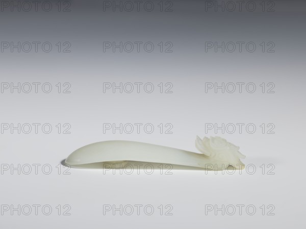 Belt clasp, Chinese, dragons iconography, Qing 1644–1911. Head in the shape of a one-horned dragon's head with a curved muzzle. Smooth, rounded handle. Milky-white jadeite, slightly translucent. Full-sculpture carved head, engraved. 17th–18th century, China. Belt buckle. Height 1.7 cm; width 9.4 cm; depth 2.1 cm., A smooth, pale white elongated object rests horizontally on a light surface, with a rounded, curved body that tapers slightly toward one end where a carved floral or petal-like decorative element sits; the carving shows layered, petal shapes with gentle ridges and the object casts a soft shadow beneath it against a background that transitions from light gray near the surface to darker gray at the top.
