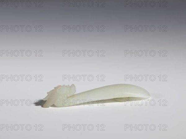 Belt clasp. Head in the shape of a one-horned dragon's head with a curved muzzle. Smooth, rounded handle. Milky-white jadeite, slightly translucent. Full-sculpture carved head, engraved. 17th–18th century, Qing 1644–1911, China. Dragons iconography. Belt buckle. Height 1.7 cm; width 9.4 cm; depth 2.1 cm., A smooth, pale off-white object rests on a flat surface against a gradient gray background; the object has an elongated, gently curved form with one end carved into an ornate sculptural head featuring layered, petal-like and curved motifs, while the opposite end tapers to a smooth rounded tip; the surface shows subtle variations in translucency and a few faint darker specks, and there is a soft shadow beneath and slightly to one side of the object; visible colors include off-white, pale cream, gray, and touches of darker gray in the shadow.