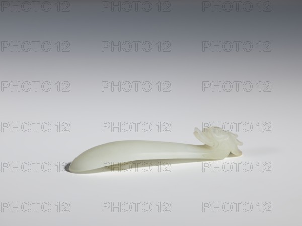 Belt clasp, Chinese, dragons iconography, Qing 1644–1911. Head in the shape of a one-horned dragon's head with a curved muzzle. Smooth, rounded handle. Milky-white jadeite, slightly translucent. Full-sculpture carved head, engraved. 17th–18th century, China. Belt buckle; height 1.7 cm, width 9.4 cm, depth 2.1 cm., A smooth, pale off-white object with a long, curved handle and a carved ornamental end resembling layered petals or a stylized floral motif rests on a light surface; the object casts a soft shadow beneath it and the background transitions from light gray near the surface to darker gray toward the top, with visible colors including off-white, light gray, and darker gray.